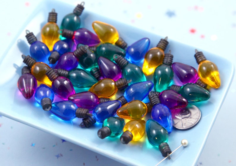 May include: Assortment of miniature plastic Christmas light bulbs in various colors including purple, teal, blue, and yellow.  These small, colorful light bulbs are ideal for crafting and miniature projects. The bulbs have dark metal caps and are translucent, allowing light to pass through.  They are displayed on a light blue tray.