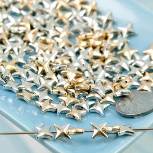 May include: A collection of star-shaped beads in gold and silver tones, scattered on a light blue tray. A few beads are strung on a needle, showcasing their design. A coin is also present, likely for size reference.