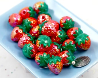 May include: A collection of red and green strawberry-shaped beads with iridescent finishes, displayed on a light blue square plate. The beads have green stems and are next to a US quarter for size comparison. These beads are ideal for jewelry making.