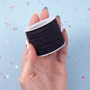 May include: A spool of black elastic cord held in a hand. The cord is wound around a white plastic spool.