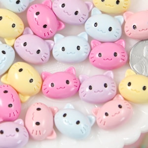 May include: A selection of pastel-coloured cat-shaped embellishments. The cat faces are in shades of pink, yellow, and blue, each with black eyes and a smiling mouth. These small, decorative pieces are ideal for crafting and adding a cute touch to various projects.