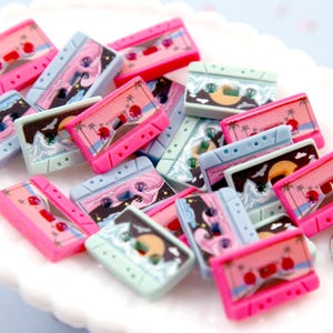 May include: A collection of colorful cassette tape shaped buttons with various beach-themed designs. The buttons are pink, blue, and green with images of palm trees, waves, and sunsets.