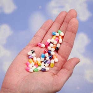 Puede incluir: Un pu&ntilde;ado de p&iacute;ldoras de c&aacute;psulas coloridas, cada una con un cuerpo blanco y una tapa de color. Las tapas son de varios colores, incluyendo rosa, verde, azul, amarillo y negro. Las p&iacute;ldoras se sostienen en una palma abierta sobre un fondo azul nublado.