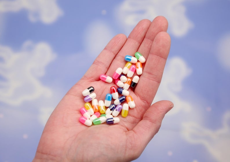 Puede incluir: Un pu&ntilde;ado de p&iacute;ldoras de c&aacute;psulas coloridas, cada una con un cuerpo blanco y una tapa de color. Las tapas son de varios colores, incluyendo rosa, verde, azul, amarillo y negro. Las p&iacute;ldoras se sostienen en una palma abierta sobre un fondo azul nublado.