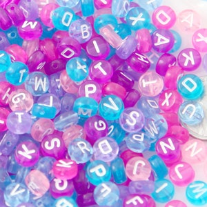 May include: A close-up of a pile of colorful plastic alphabet beads. The beads are translucent and have white letters on them. The beads are in shades of pink, purple, and blue.