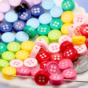 May include: A collection of colorful, round buttons in various shades of blue, green, yellow, pink, and red. Each button has four small holes and is displayed on a white, scalloped dish. A needle is inserted through some of the buttons.