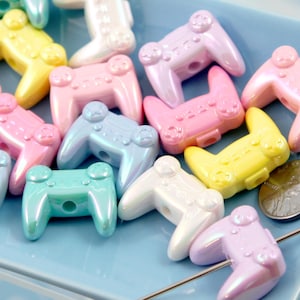 May include: A selection of iridescent game controller beads in pastel colours, including pink, blue, yellow, and white. The beads are displayed on a light blue surface, suggesting a craft or jewellery-making theme.