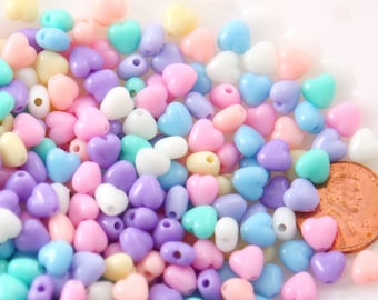 May include: A collection of pastel-colored heart-shaped beads in shades of pink, purple, blue, yellow, and white. The beads are scattered, with a penny for size comparison. These beads are ideal for jewelry making and crafting.