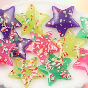 May include: A collection of colorful star-shaped resin embellishments. The stars are in shades of purple, green, pink, and yellow, with a confetti-like pattern of red, white, and green. These decorative stars are ideal for crafting and embellishing various projects.
