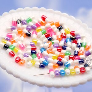 Puede incluir: Un conjunto de cuentas en forma de c&aacute;psula de colores, incluyendo rojo, azul, amarillo y morado, se encuentran dispersas sobre un plato blanco con borde festoneado. Se incluye una moneda para referencia de tama&ntilde;o. El fondo es azul claro.