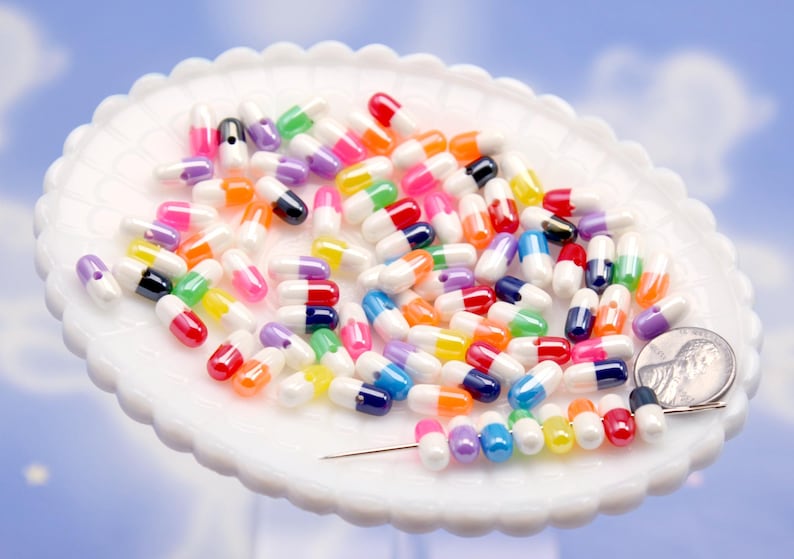 Puede incluir: Un conjunto de cuentas en forma de c&aacute;psula de colores, incluyendo rojo, azul, amarillo y morado, se encuentran dispersas sobre un plato blanco con borde festoneado. Se incluye una moneda para referencia de tama&ntilde;o. El fondo es azul claro.