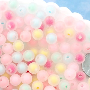May include: A selection of pastel-coloured round beads in pink, yellow, and green. The beads are scattered in a white, scalloped-edge dish. A penny is included for size reference. These beads are suitable for jewellery making and crafts.