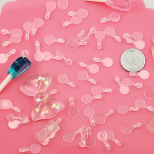 May include: A collection of clear plastic charms in various shapes, including circles, stars, and bows. The charms are scattered on a pink surface, along with a blue and white lollipop and a silver coin.