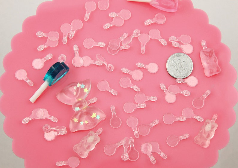 May include: A collection of clear plastic charms in various shapes, including circles, stars, and bows. The charms are scattered on a pink surface, along with a blue and white lollipop and a silver coin.