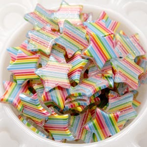 以下が含まれることがあります： 虹色のストライプの星形の装飾が詰まったボウル。星は光沢のあるキラキラした素材でできています。