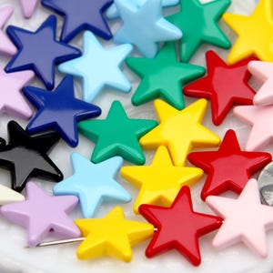 May include: A collection of multi-colored star-shaped beads in shades of red, yellow, green, blue, black, white, and pink. A silver-colored coin is positioned on the right side for scale, showing the relative size of the star beads.