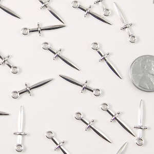 May include: Silver dagger charms with a loop for jewellery making. The charms are scattered on a white surface, with a penny for size comparison. The daggers have a pointed blade and a crossguard.
