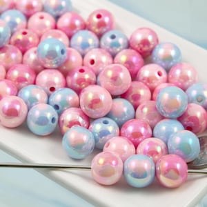 Könnte beinhalten: Eine Sammlung von irisierenden Perlen in Rosa und Blau auf einer weißen Oberfläche. Die Perlen sind rund und glänzend. Eine Nadel liegt daneben, was auf Bastelarbeiten oder Schmuck hindeutet.