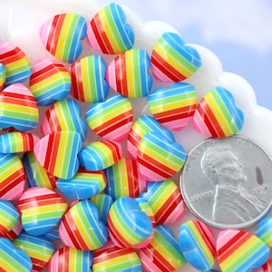 以下が含まれることがあります： 虹色のストライプのハート型のレジンビーズの山をクローズアップした写真。ビーズはアメリカの25セント硬貨と同じくらいの大きさです。