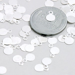 May include: A large quantity of silver-toned metal round charms with a small loop on the top. The charms are scattered on a white surface next to a US one cent coin.