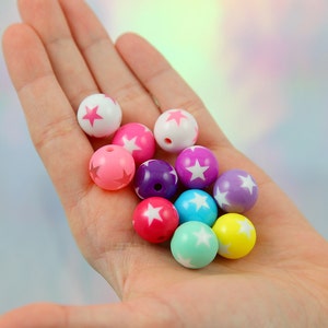 May include: A selection of colourful beads, each with a star pattern. The beads are in shades of pink, purple, blue, and yellow. The round beads are held in a hand. Each bead has a white star design.