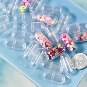 May include: Clear plastic capsules, some filled with colorful beads, hearts, and other small decorative items, are scattered on a light blue surface. A US penny is included for size reference. The capsules are empty and ready to be filled.