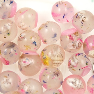 May include: A collection of clear, spherical beads with embedded seashells and colorful floral accents. The beads have a pink hue and are arranged on a white surface. A US quarter is visible for size comparison.