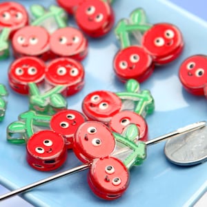 May include: A collection of red cherry-shaped beads with smiling faces and green stems, arranged on a light blue surface. A silver needle and a penny are also present. The beads are likely for crafting or jewelry making.