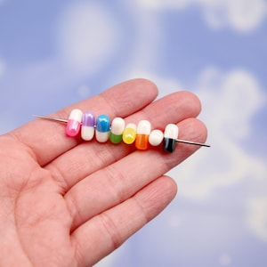 Puede incluir: Una colecci&oacute;n de cuentas coloridas en forma de c&aacute;psula ensartadas en una aguja plateada. Las cuentas son de varios colores, incluyendo rosa, morado, azul, verde, amarillo, naranja y negro. El fondo es azul suave con formas de nubes blancas.
