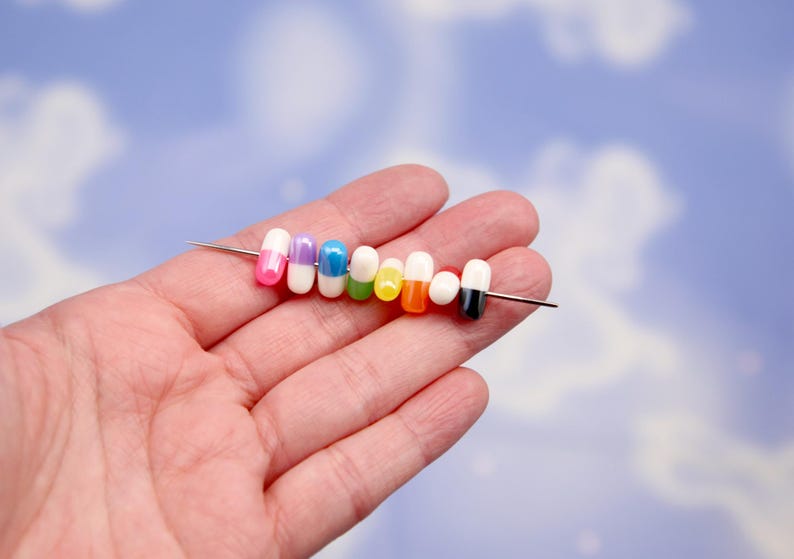 Puede incluir: Una colecci&oacute;n de cuentas coloridas en forma de c&aacute;psula ensartadas en una aguja plateada. Las cuentas son de varios colores, incluyendo rosa, morado, azul, verde, amarillo, naranja y negro. El fondo es azul suave con formas de nubes blancas.