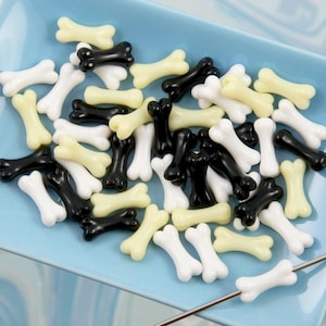 May include: A light blue square dish filled with bone-shaped beads in black, white, and pale yellow. A metal needle with a bone bead is in the foreground. The beads are likely for jewelry making or crafts.