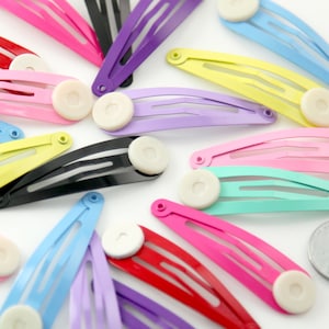 May include: A selection of brightly coloured metal hair slides in shades of pink, purple, yellow, blue, and black. Each slide has a round, off-white accent. The slides are arranged on a light background.