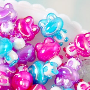 May include: A collection of iridescent frog-shaped beads in shades of pink, purple, and blue. The beads have a glossy finish and are perfect for crafting jewelry or other projects.