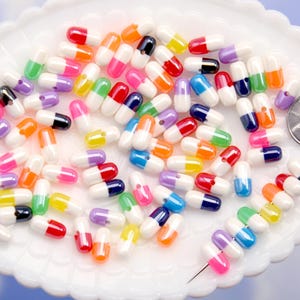Puede incluir: Una colecci&oacute;n de cuentas coloridas en forma de c&aacute;psula en varios colores, incluyendo rojo, naranja, amarillo, verde, azul, rosa, morado y negro, est&aacute;n dispersas sobre un plato blanco con borde festoneado. Se incluye una moneda como referencia de tama&ntilde;o.