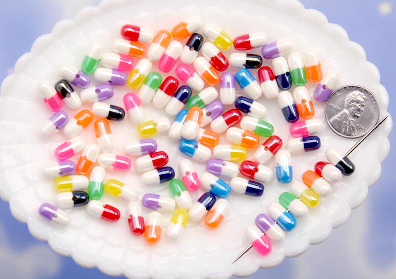 Puede incluir: Una colecci&oacute;n de cuentas coloridas en forma de c&aacute;psula en varios colores, incluyendo rojo, naranja, amarillo, verde, azul, rosa, morado y negro, est&aacute;n dispersas sobre un plato blanco con borde festoneado. Se incluye una moneda como referencia de tama&ntilde;o.
