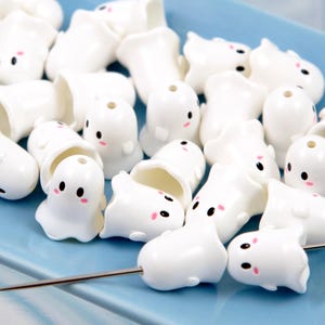 May include: A collection of white ceramic ghost-shaped beads with black eyes and pink cheeks, scattered on a light blue tray. A needle is placed among the beads. The beads have a hole at the top, suggesting they are for jewelry making.