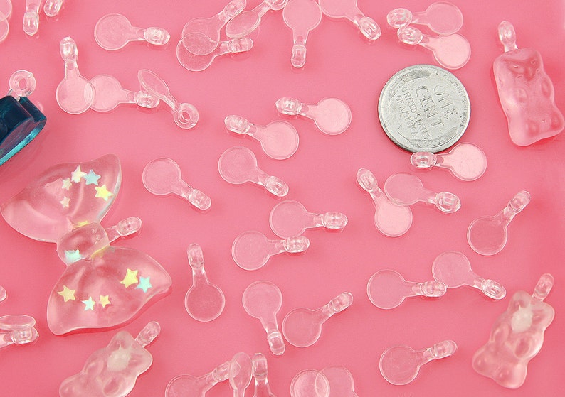 May include: A collection of clear plastic charms in various shapes, including circles, stars, and bows. The charms are scattered on a pink surface. There is also a US coin with the text "One Cent" visible in the image.