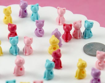 May include: A collection of colorful plastic cat-shaped beads. The beads are in various colors, including pink, blue, yellow, and purple. They are all about 1 inch tall and have a hole in the center for stringing.