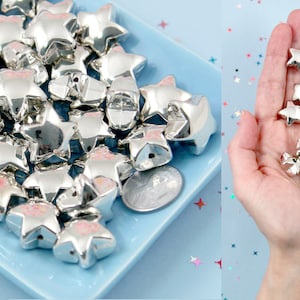 May include: A large pile of silver star-shaped beads on a light blue surface. The beads are about 1 inch in diameter and have a shiny, metallic finish.