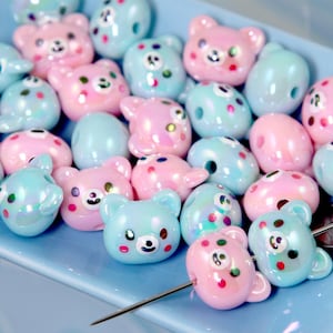 May include: A collection of pastel-colored beads in a blue dish. The beads are shaped like bears and other rounded forms, with iridescent finishes and painted details. A needle is inserted through one of the beads.