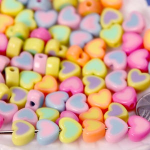 May include: A collection of small, heart-shaped beads in pastel shades of pink, yellow, blue, orange, and purple. A silver needle is threaded through five beads at the bottom, and a US penny is visible for scale next to the pile of loose beads.