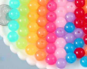 Puede incluir: Una colección de cuentas redondas y coloridas en un arcoíris de tonos, incluyendo turquesa, amarillo, naranja, rosa, morado y rojo. Las cuentas están dispuestas sobre una superficie blanca y festoneada. Se incluye una moneda como referencia de tamaño.