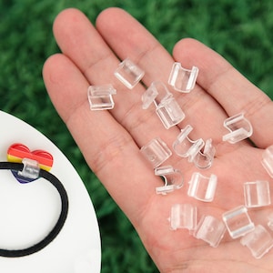 May include: A collection of clear plastic hair tie clips, some held in a hand, and a black hair tie with a rainbow heart-shaped charm. The clips are small, rectangular, and have a U-shaped opening.