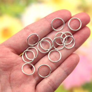 Op de afbeelding: Een hand die een stapel zilveren springringen vasthoudt. De ringen zijn klein en rond en perfect voor het maken van sieraden of andere knutselprojecten.