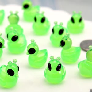 Op de afbeelding: Een verzameling kleine, doorschijnende groene alien-eendenbeeldjes met zwarte ogen en antennes. De figuren zijn gerangschikt op een wit, geschulpte oppervlak, wat een speelse scène creëert. Er is ook een munt aanwezig.