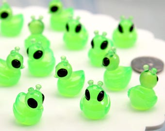 May include: A collection of small, translucent green alien duck figurines with black eyes and antennae. The figures are arranged on a white, scalloped surface, creating a playful and whimsical scene. A coin is also present.