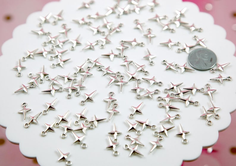 Puede incluir: Una gran cantidad de abalorios en forma de estrella de color plateado con dos anillos de salto en cada abalorio. Los abalorios est&aacute;n esparcidos sobre una superficie blanca.