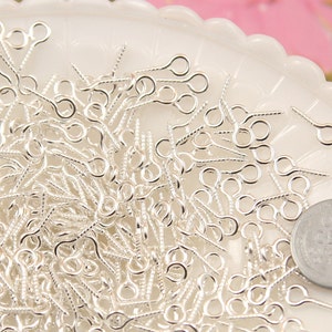 May include: A close-up image of a pile of silver-toned eye pins. The eye pins are small and have a loop at one end and a pointed end. The eye pins are arranged in a random pattern.
