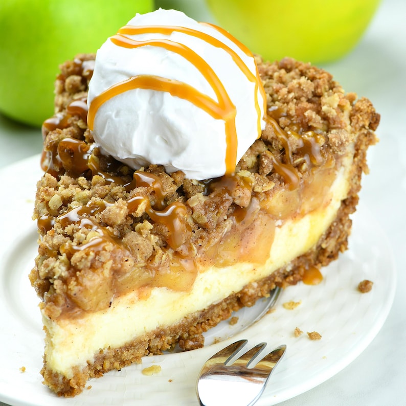 Delicious slice of apple crisp cheesecake pie. It has a crumbly streusel topping, layered over spiced apples and creamy cheesecake. The slice is topped with whipped cream and drizzled with caramel sauce, making it look indulgent and mouth-watering.