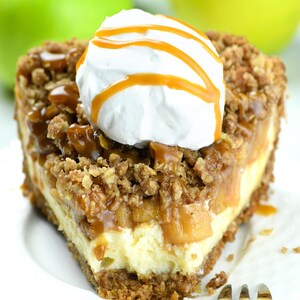 Slice of apple crisp cheesecake pie with a bite taken from it. It features a crumbly oat topping over baked apples and creamy cheesecake filling. The dessert is topped with a dollop of whipped cream and a caramel drizzle, all served on a white plate.